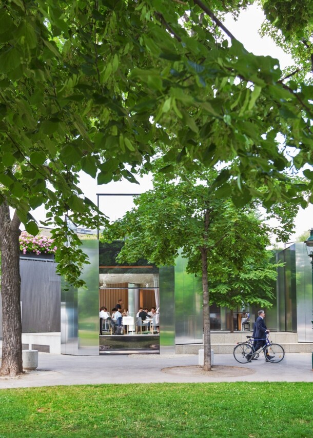 Rendering des Wiener Stadtparks mit Bäumen und Wegen, im Hintergrund ein Restaurant mit offenen Türen und Gästen im Inneren