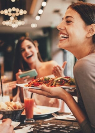 Eine Gruppe fröhlicher Personen in einem Restaurant. Eine lachende Frau hält einen Teller in der Hand.
