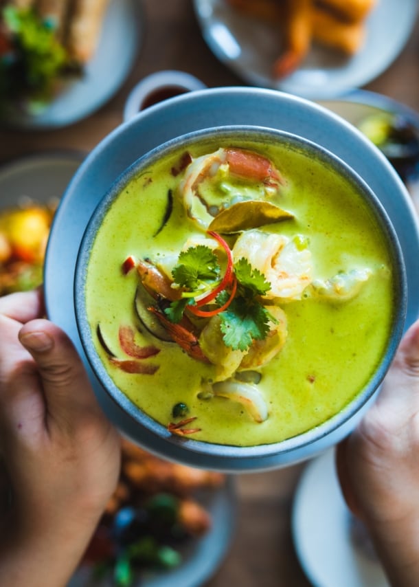 Zwei Hände halten eine Schale mit einem grünen Curry-Gericht über einen gedeckten Tisch mit vielen weiteren Speisen.