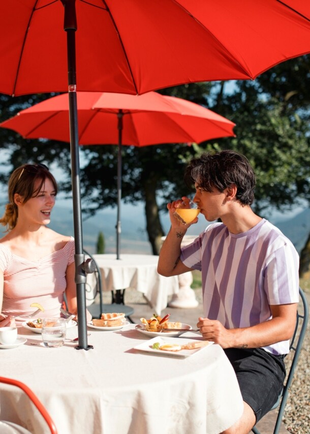 Ein Paar frühstückt auf einer Terrasse an einem Tisch bei Sonnenschein, im Hintergrund eine Hügellandschaft.
