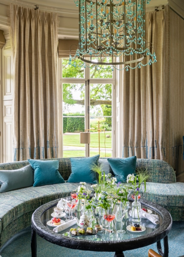 Luxuriöser Salon mit Kronleuchter und geschwungenem Sofa vor Panoramafenstern mit Blick in einen Garten.