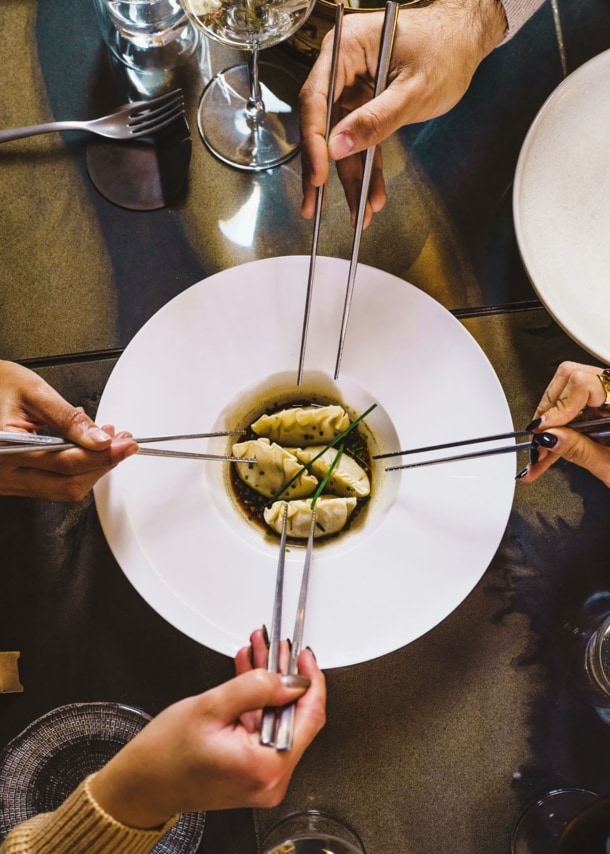 Aufsicht eines gedeckten Restauranttisches mit einem weißen Teller mit Teigtaschen, umgeben von vier Händen mit Essstäbchen.
