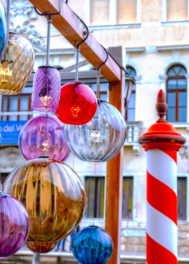 Nahaufnahme mehrerer, hängender Murano-Glaslichter, im Hintergrund eine venezianische Häuserfront.