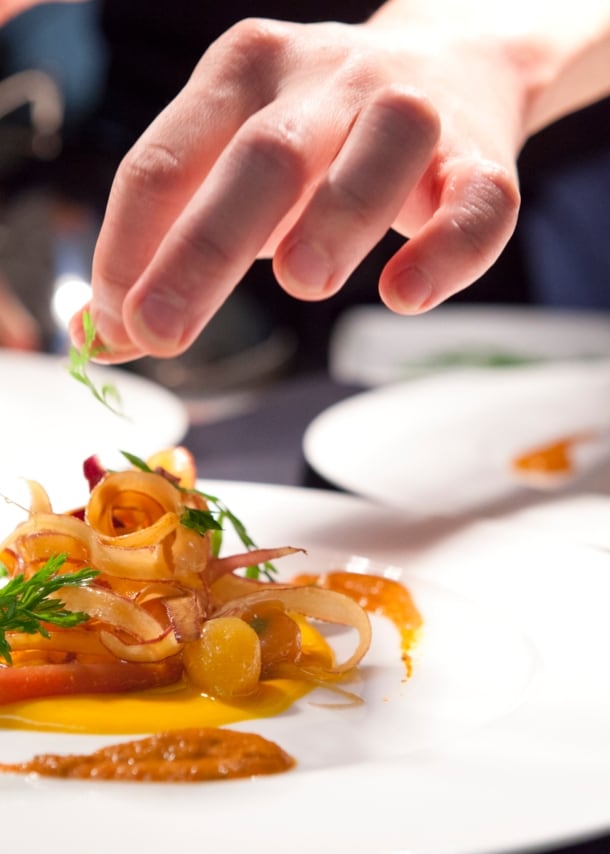 Eine Hand garniert ein Gericht auf einem weißen Teller in einer Restaurantküche.