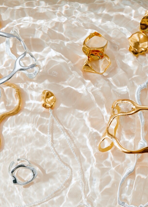 Unterschiedliche Schmuckstücke in Gold und Silber liegen auf Wasser mit Lichtspiel