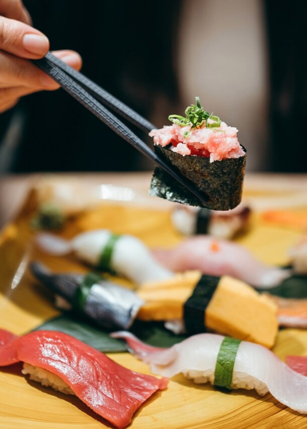 Eine Person hält eine Rolle Maki Sushi zwischen zwei Essstäbchen über einer Bambusplatte mit Nigiri auf einem Tisch in einem Restaurant.