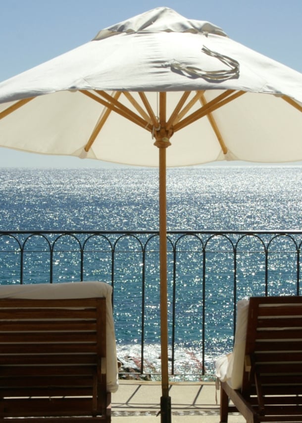 Terrasse und Geländer mit Blick aufs Meer, auf der mehrere Liegestühle und ein weißer Sonnenschirm stehen.