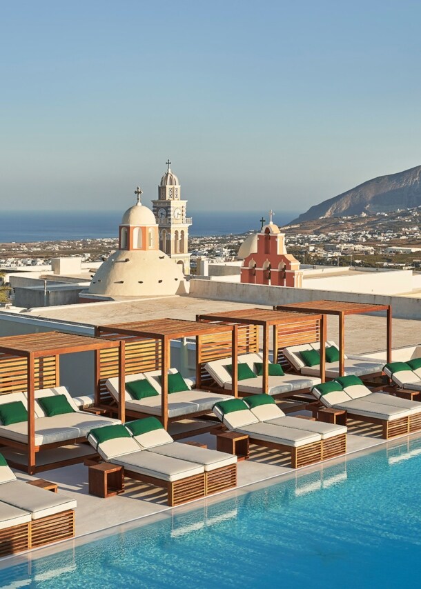 Dachterrasse mit zahlreichen Sonnenliegen am Infinity-Pool auf Santorini, im Hintergrund Meeresküste.