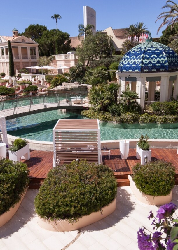 Ein lang gestreckter, mit Palmen, Blumen und Daybeds gesäumter Pool, über den zwei Brücken führen.