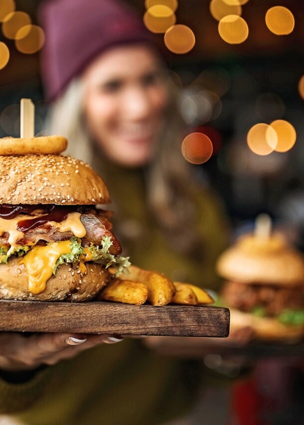 Ein Burger auf einem Brett, gehalten von einer Frau in Unschärfe in einem Lokal.