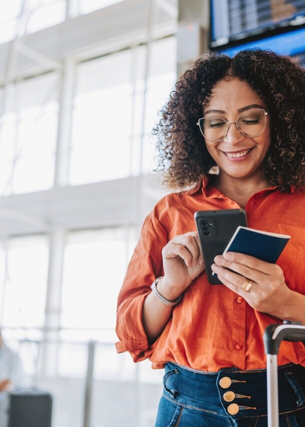 ine Frau bedient lächelnd ihr Smartphone in einem Flughafenterminal, in ihrer Hand ein Reisepass.