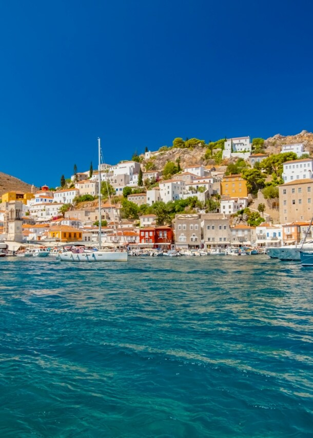 Mediterrane Ortschaft in Hügellage mit Hafenpromenade, im Vordergrund türkisblaues Meer mit Booten.