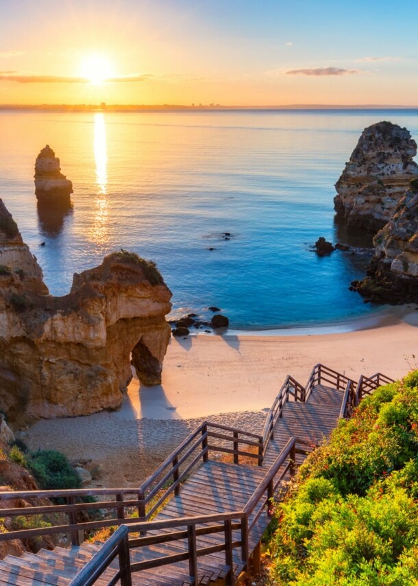 Der Strand Praia do Camilo in Portugal bei Sonnenuntergang, zu dem über Felsen eine Holztreppe nach unten führt.