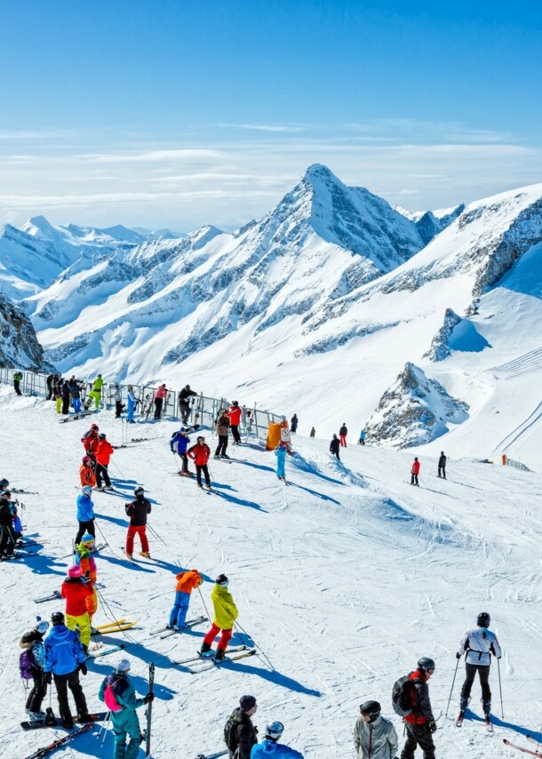 Skifahrende auf einem Plateau auf einer Skipiste vor schneebedecktem Gipfelpanorama.