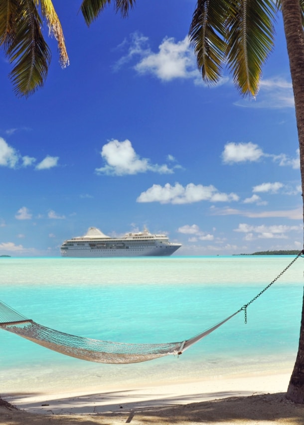 Ein Kreuzfahrtschiff auf türkisblauem Meer, umrahmt von zwei Palmen mit Hängematte an einem Sandstrand im Vordergrund.