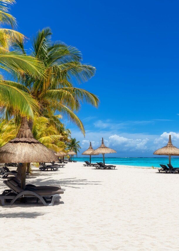 Weißer Sandstrand auf Mauritius mit Sonnenliegen, Strohschirmen, grünen Palmen und türkisfarbenem Meer.