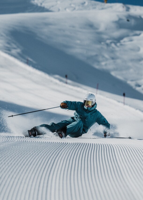 Ein Skifahrer mit Helm im türkisen Schneeanzug bei der Abfahrt auf einer Piste.