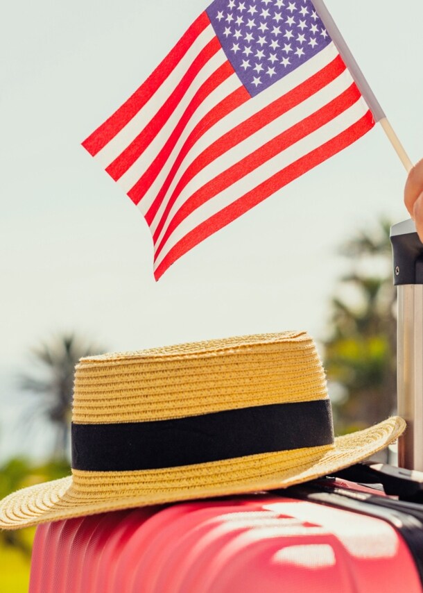 Ein Strohhut auf einem pinken Hartschalenkoffer, eine Hand am Griff hält eine kleine US-amerikanische Flagge, im Hintergrund Plamen und ein Flugzeug am Himmel.