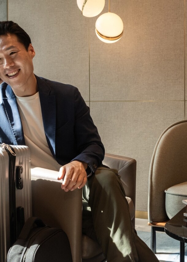 Ein fröhlicher asiatischer Geschäftsmann sitzt im Sessel einer modernen Flughafen-Lounge und telefoniert an seinem Smartphone, neben ihm steht ein großer Reisekoffer.