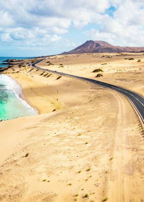 Ein weißes Auto fährt auf einer Schnellstraße durch eine Wüstenlandschaft am türkisblauen Meer.