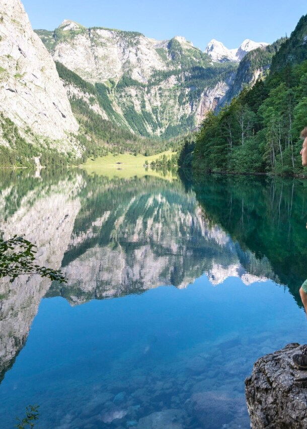 Eine Person mit Wanderrucksack steht auf einem Stein vor dem Königssee mit Blick auf die Berge.