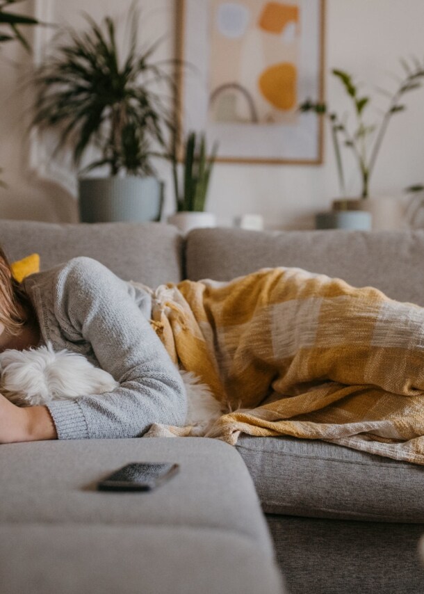 Eine Frau liegt schlafend mit einer Decke auf einer grauen Couch, im Arm hält sie einen Hund.