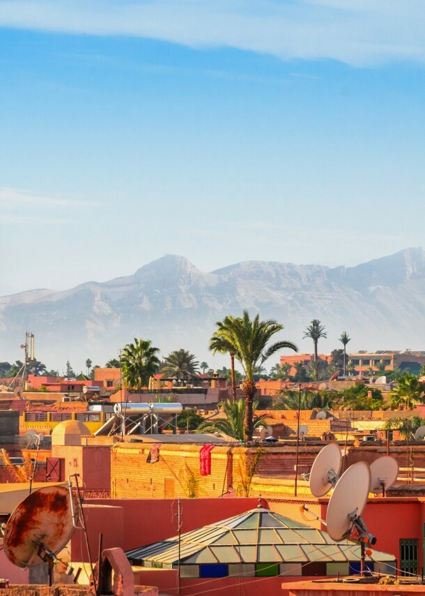 Stadtpanorama von Marrakesch mit Minarett vor Gebirgskette im Hintergrund unter blauem Himmel.