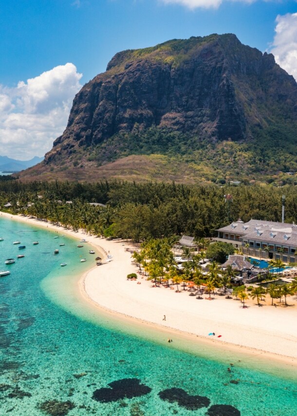 Küstenpanorama mit Hotelresort an einem palmengesäumten, weißen Sandstrand an türkisblauem Meer, im Hintergrund Berge.