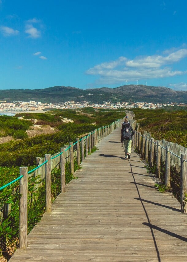 Eine unkenntliche Person beim Wandern in Portugal auf einem Holzsteg, der in Küstennähe über grüne Vegetation führt.