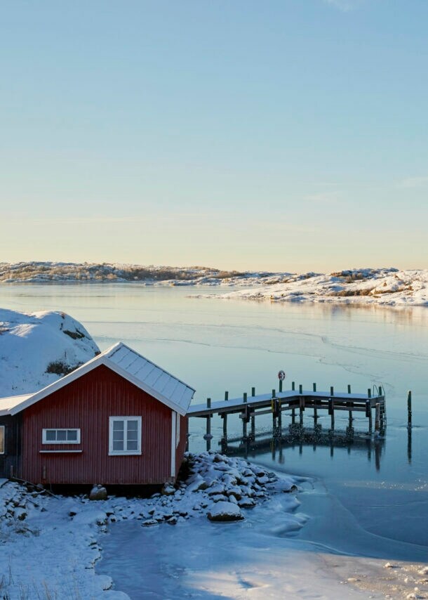 Ein rotes Holzhaus mit Steg an einem zugefrorenen See in verschneiter Landschaft in Schweden.