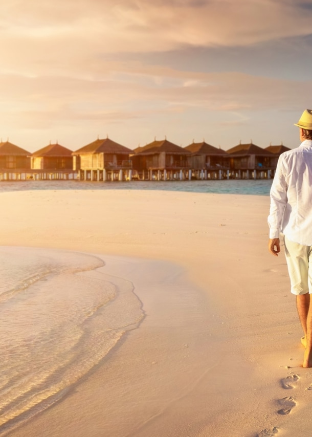 Rückansicht eines händchenhaltenden Paares in heller Sommerkleidung und mit Strohhüten an einem Strand vor einem Holzstegmit Wasserbungalows.