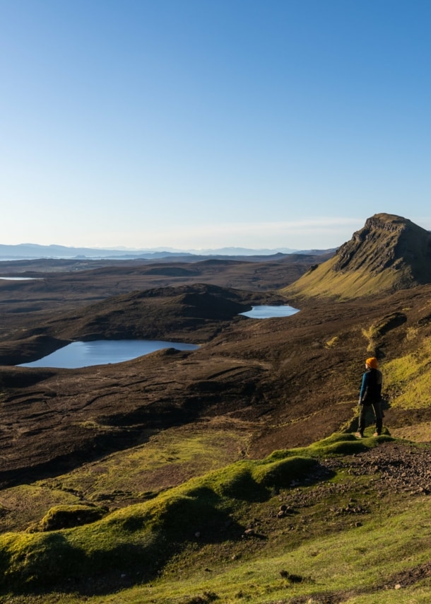 Eine Person steht in einer Landschaft aus Wiesen, Hügeln und Seen in Schottland. 