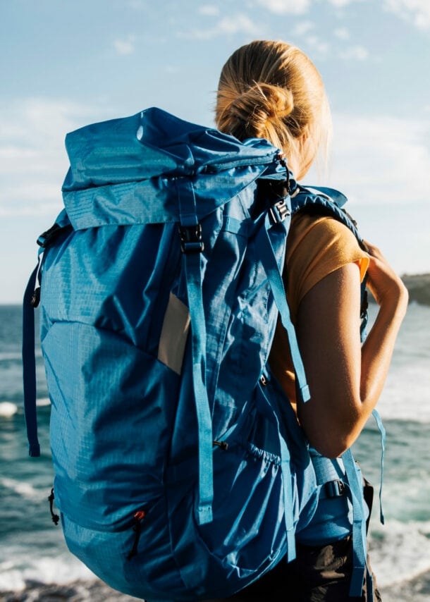 Rückansicht einer Frau mit großem blauen Rucksack am Strand, die aufs Meer blickt.