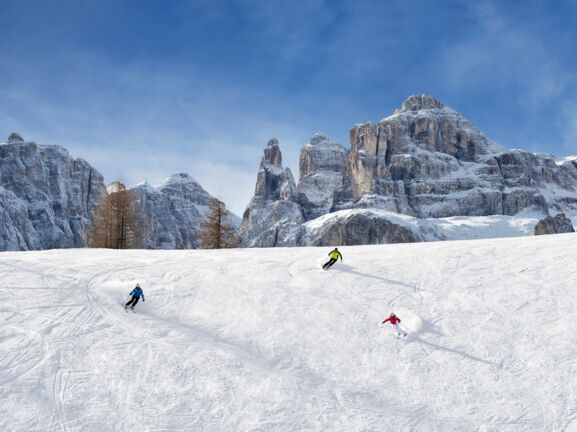 Panorama einer Skipiste mit drei Skifahrern vor markanten Felsformationen eines Bergmassivs