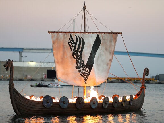 Aufnahme eines brennenden Wikingerschiffs auf dem Meer mit einem Segel, das das Logo der „Vikings“-Serie zeigt