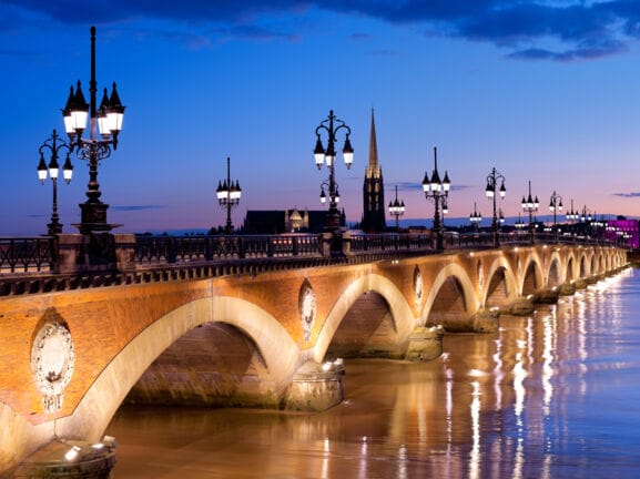 Eine alte Steinbrücke mit erleuchteten Straßenlaternen verläuft über einen Fluss in Bordeaux bei Nacht