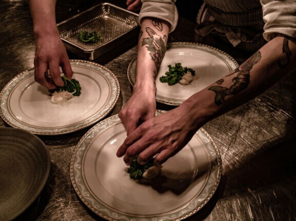 Nahaufnahme von Händen beim Anrichten von Speisen auf großen, weißen Tellern in einer Restaurantküche.