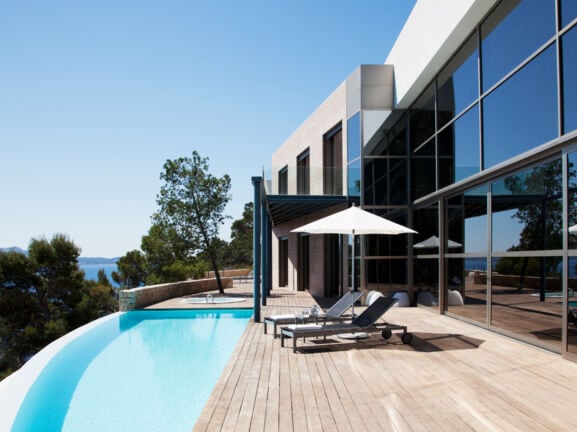 Ein Haus mit großer Glasfensterfront und angeschlossener Holzterrasse mit Pool. Auf der Terrasse befinden sich ein Sonnenschirm und zwei Liegen.