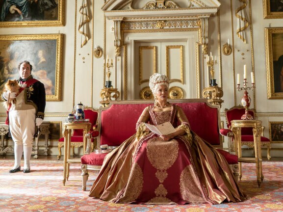 Eine Königin sitzt auf einem roten Samtsofa in einem Palast mit weiß-goldenem Dekor.