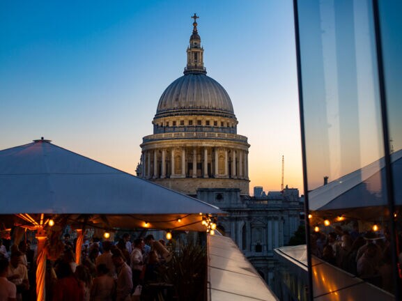 Belebtes Restaurant auf Dachterrasse vor der Kuppel der St Paul’s Kathedrale bei Abenddämmerung.