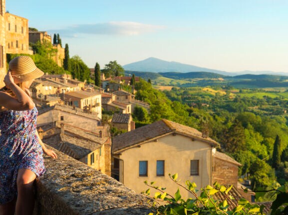 Eine Frau im Sommerkleid blickt von einer Mauer in einem italienischen Dorf hinunter auf eine grüne Hügellandschaft bei warmem Sonnenlicht.