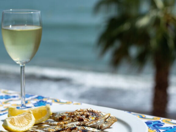 Gegrillte Sardinen auf einem weißen Teller neben einem Glas Weißwein, dahinter Meer mit Palme in Unschärfe.