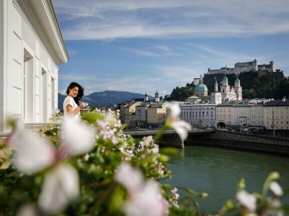 Eine Frau steht mit Kaffeetasse auf einem Balkon am Fluss, ihr gegenüber die Salzburger Altstadt mit Burgfestung.