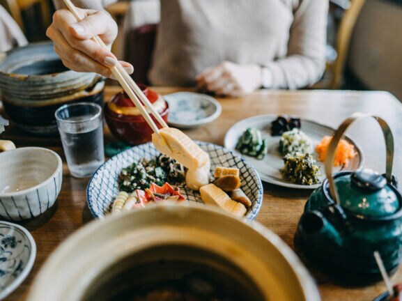 Auf einem Tisch stehen mehrere Schälchen mit verschiedenen asiatischen Gerichten. Eine unkenntliche Person hält mit Stäbchen etwas zu essen hoch.