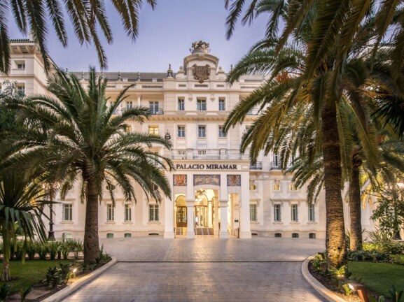 Imposante, helle Frontfassade eines Grandhotels mit Vorplatz, im Vordergrund Begrünung mit Palmen.