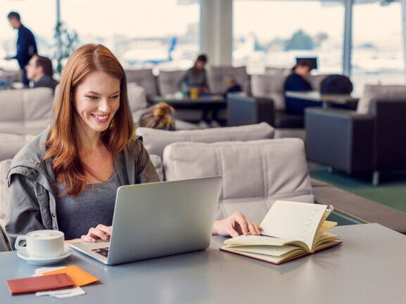 Eine lächelnde Frau arbeitet an ihrem Laptop an einem Tisch in einer Flughafen-Lounge.