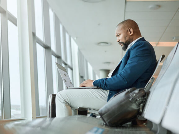 Ein Mann, der am Flughafen am Fenster sitzt und einen aufgeklappten Laptop auf dem Schoß hat.