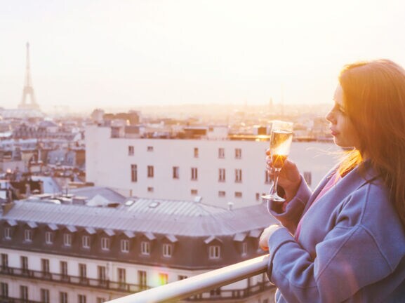 Eine Frau, die bei Sonnenuntergang in Paris auf einem Balkon steht und ein Glas Sekt in der Hand hält.