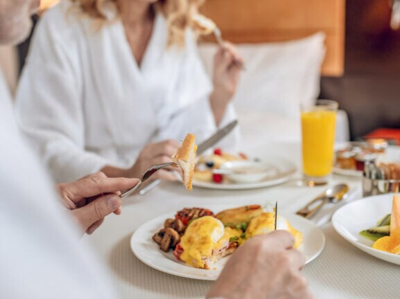 Nahaufnahme von zwei gesichtslosen Personen in weißen Bademänteln, die an einem eingedeckten Tisch in einem Hotelzimmer frühstücken.