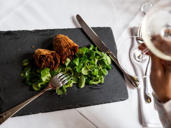 Aufsicht eines panierten Fleischgerichtes an Feldsalat auf einer Schieferplatte auf einem edel gedeckten Tisch, im Vordergrund eine Hand mit einem Glas Weißwein.